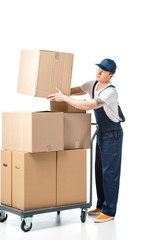 handsome mover in uniform transporting cardboard box near hand truck with packages isolated on white