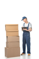 partial view of handsome mover in uniform writing in clipboard near cardboard boxes isolated on white