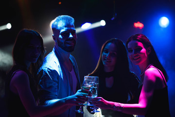  Young friends dancing with glasses of champagne in hands. Against lighting devices as background. Young people's friends are dancing.