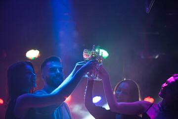  Young friends dancing with glasses of champagne in hands. Against lighting devices as background. Young people's friends are dancing.