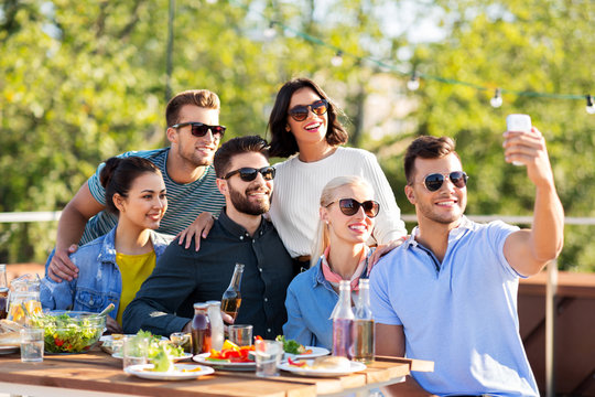 leisure and people concept - happy friends with non alcoholic drinks having dinner party on rooftop in summer and taking selfie by smartphone