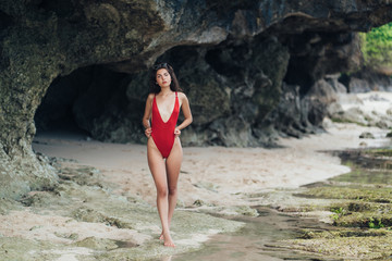 Slender tanned girl in swimwear and sunglasses posing near stone at rocky beach by ocean.