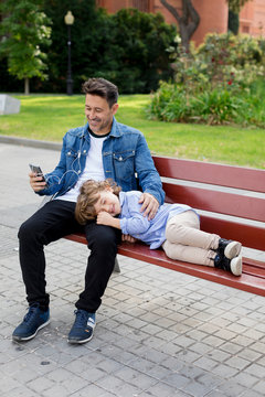 Happy father with son on a bench using cell phone and earbuds