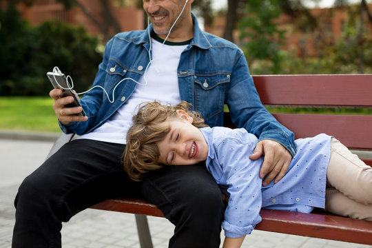 Happy father with son on a bench using cell phone and earbuds