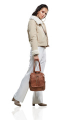Full length side shot of lady, wearing short Afghan coat and white trousers. The girl is making a step, tilting head and holding shoulder strap of brown rucksack with flap top, adorned with zippers.