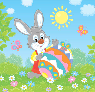 Little Easter Bunny and a colorfully decorated big egg among flowers and flittering butterflies on a green lawn on a sunny spring day, vector illustration in a cartoon style