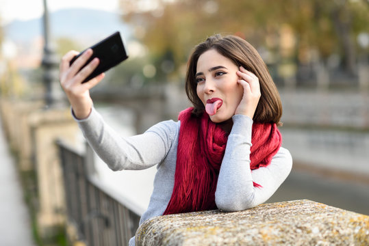 Portrait of young woman wearing red scarf sticking out tongue taking selfie with cell phone