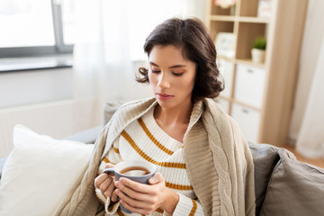 health, cold and people concept - sad sick young woman in blanket sitting on sofa and drinking hot tea at home