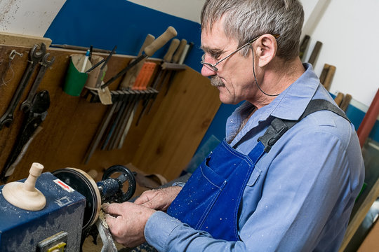 A white elderly man with glasses works at a lathe in a carpentry workshop. The concept of increasing the retirement age