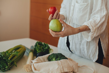 Female hands holding green fresh apple after doing shopping in the local farm supermarket, putting on the table vegetables and fruit from a cotton shopper bag instead of a plastic bag