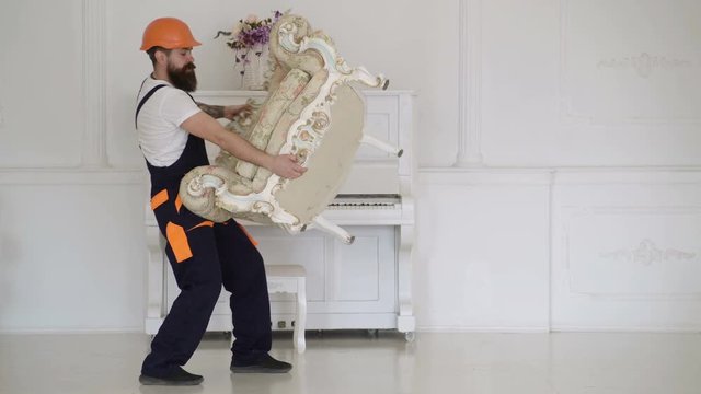 Loader moves armchair. Heavy loads concept. Man with beard worker in helmet and overalls lifts up, efforts to move armchair, white background. Courier delivers furniture, move out, relocation.