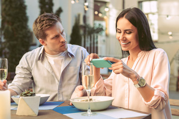 Beaming dark-haired woman in light blouse photographing her dinner