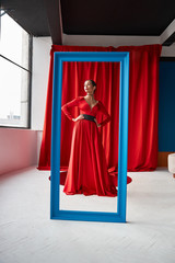 Fototapeta premium Young sexy woman in long red dress standing behind blue frame. Red curtains on background.