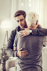Twenty-years-old short-haired kind man hugging ill upset old grey-haired relative