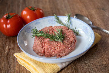 Fresh raw minced meat with rosemary on a plate and tomato on wooden table
