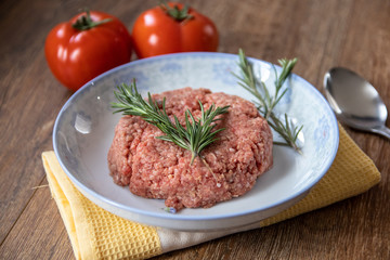 Fresh raw minced meat with rosemary on a plate and tomato on wooden table