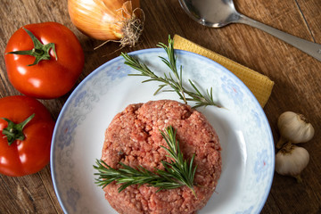 Fresh raw minced meat with rosemary on a plate and tomato, garlic and onion on wooden table