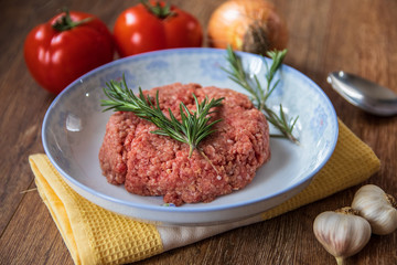 Fresh raw minced meat with rosemary on a plate and tomato, garlic and onion on wooden table