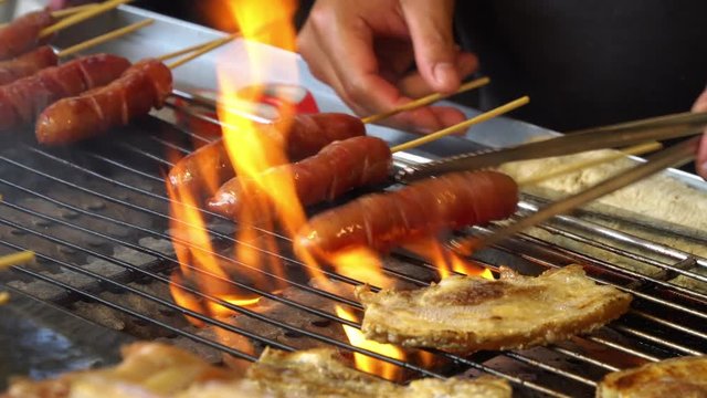 4K Close up shot of a men hand use stainless steel tongs cooking grilled sausage and streaky pork steak on fire. Grill barbecue meat on iron sieve. Street food in Taipei, Taiwan.