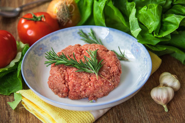 Fresh raw minced meat with rosemary on a plate and tomato, garlic, lettuce and onion on wooden table. Composition with meat and fresh organic vegetable.