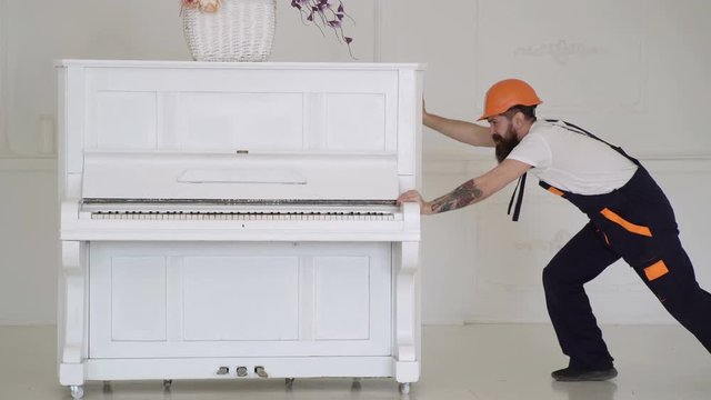 Man with beard worker in helmet and overalls pushes, efforts to move piano. Loader moves piano instrument. Man with beard, worker in overalls and helmet fall asleep tired, white background.