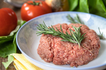 Fresh raw minced meat with rosemary on a plate and tomato, garlic, lettuce and onion on wooden table. Composition with meat and fresh organic vegetable.
