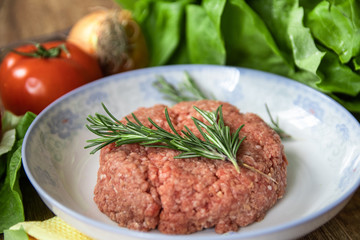 Fresh raw minced meat with rosemary on a plate and tomato, garlic, lettuce and onion on wooden table. Composition with meat and fresh organic vegetable.