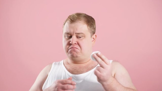 Funny glad jouful man in undershirt applying tonic by cotton pad on face isolated on pink studio background. Hygiene concept, cosmetical goods for skin care
