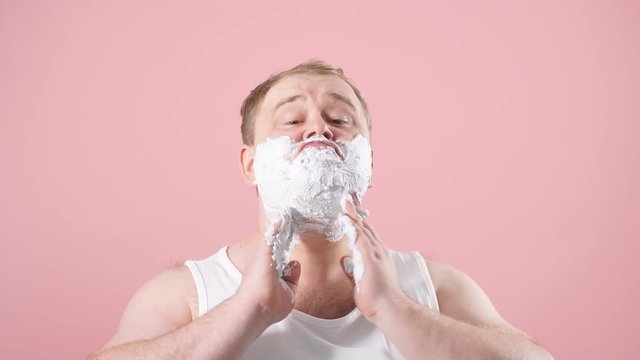 Cheerful happy man with shaving foam on his face doing morning routine procedure isolated over pink background.