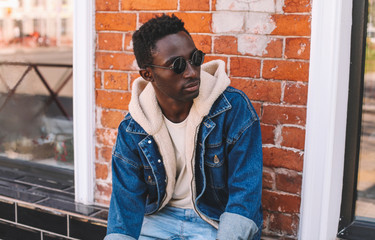 Fashion portrait african man wearing jeans jacket sitting on city street over brick textured wall background