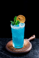 Beautiful blue cocktail with ice, decorated with mint and a slice of dry orange in a beautiful faceted glass on a round wooden board on a black background. Close up. Space. Dark background