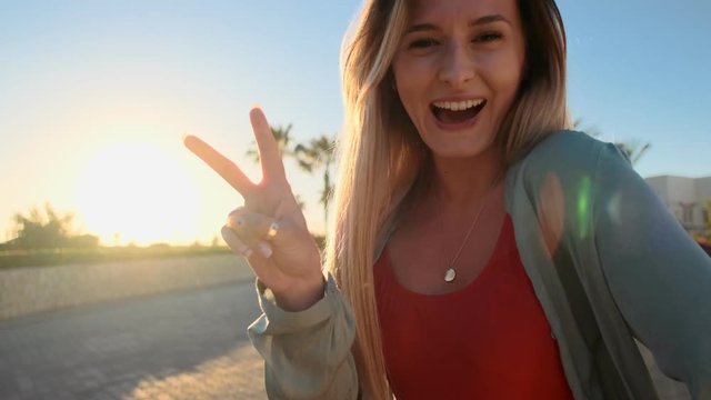 Sun portrait of attractive girl in red swimsuit looking at camera and making peace sign with hand or V sign with fingers with palmas background. Woman enjoying summer vacation on tropical resort.