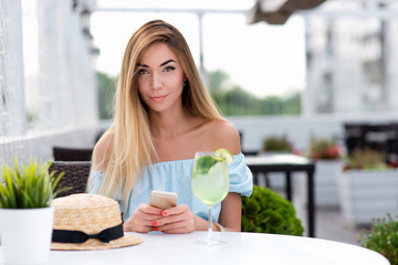 Beautiful girl with long hair. A woman summer cafe restaurant. Sits at table blue dress, close-up. In hand telephone, table glass of cocktail. Emotions relaxation enjoyment and comfort from work done.