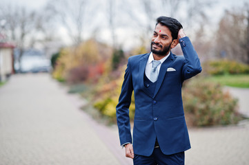 Stylish beard indian man with bindi on forehead, wear on blue suit posed outdoor.