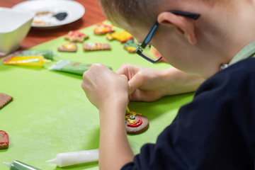 Dekorowanie cynamonowych ciastek przez dziecko. Dłonie chłopca tworzą twarz z kolorowego lukru wyciskanego z tubek. Na drugim planie ciastka nieudekorowane.