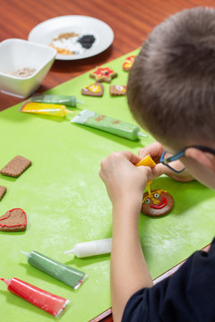 Dekorowanie cynamonowych ciastek przez dziecko. Dłonie chłopca tworzą twarz z kolorowego lukru wyciskanego z tubek. Na drugim planie ciastka nieudekorowane.