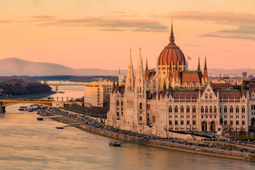 Cityscape of Budapest with bright parliament illuminated by last sunshine before sundown and Danube river with bridge. Pink and purple colors of sky reflecting in water during sunset.