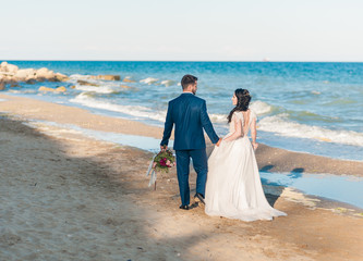 happy newlywed couple holding hands and walking around ocean. Wedding bride and groom with bouquet posing near sea in summer time