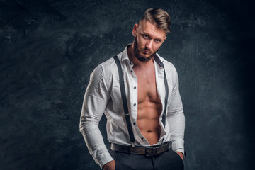 A stylish young man with a muscular body in the unbuttoned white shirt with suspenders. Studio photo against a dark wall background