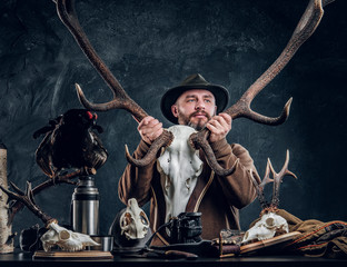 Mature bearded hunter in hat holding a big deer skull while standing next to a table with equipment...