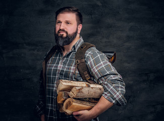 Portrait of a bearded woodcutter with a backpack dressed in a plaid shirt holding firewood. Studio photo against a dark textured wall