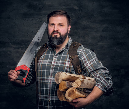 Portrait of a bearded woodcutter with a backpack dressed in a plaid shirt holding firewood and hand saw. Studio photo against a dark textured wall