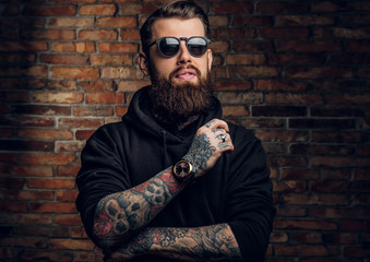 A stylish tattooed guy in a black hoodie and sunglasses. Studio photo against a brick wall