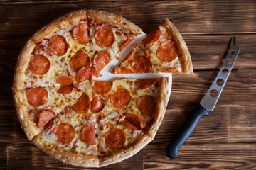 Homemade pepperoni pizza and cheese knife lie on a natural wooden surface made of pine boards. Daylight