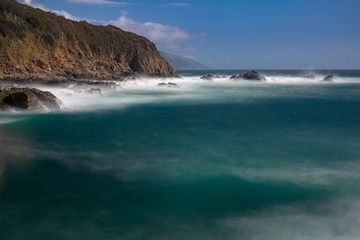 Long exposure of deep blue water, swirling surf, and rocky cliffs of central California