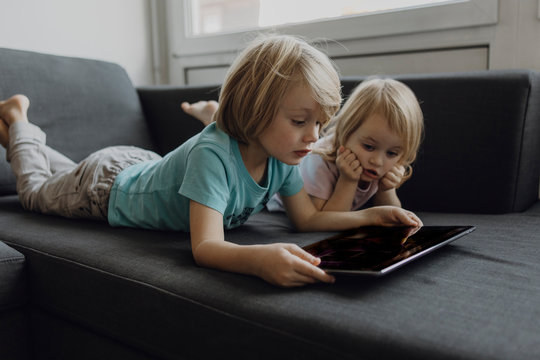 Brother and little sister lying on the couch at home using digital tablet