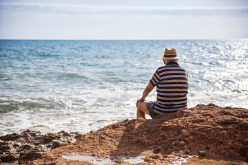 Italy, Sicily, Cava D'aliga, senior man sitting at the coast and looking to the sea