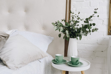 Two green cups with saucers and eucalyptus bouquet on bedside table or night stand. Modern interior decoration.