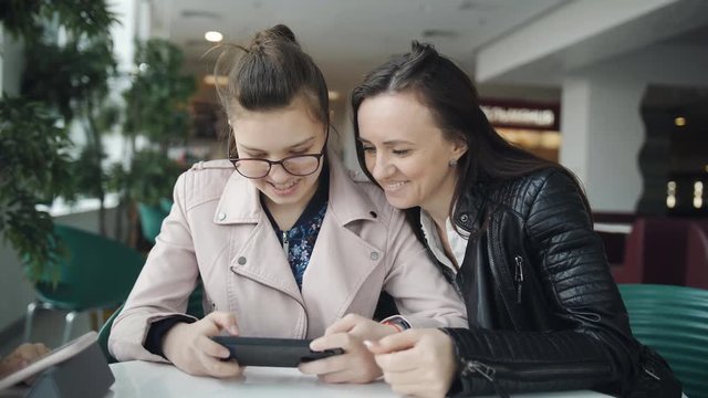 Young mother and teen daughter looking at smartphone in anticipation of an order in cafe. Beauty teen girl using smartphone showing photos to her mother. Discussing, having chat.