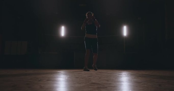 Training a beautiful female boxer doing punches in a dark room. Steadicam shot. Preparation for self-defense and fight in the ring
